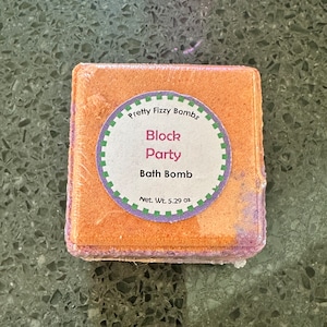 May include: A square bath bomb with orange and purple hues, wrapped in clear plastic. A circular label reads "Pretty Fizzy Bombs Block Party Bath Bomb" with a net weight of 5.29 oz.