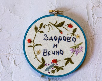 May include: Embroidered hoop art with the words "ЗДОРОВО И ВЕЧНО" in blue thread. The design features a floral wreath with green leaves, red berries, and purple and white flowers, set against a cream-colored fabric.