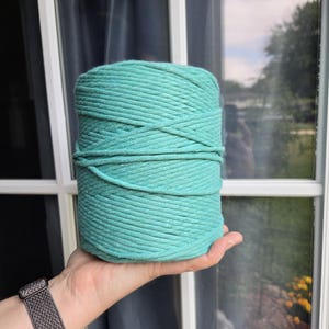 May include: A cylindrical spool of thick, teal-colored cotton cord wound tightly in a continuous spiral. The cord features a smooth, braided texture and a consistent, vibrant green-blue hue throughout the entire length of the material.