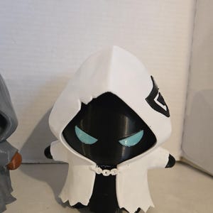 May include: A small, stylized figurine featuring a black body and a white hooded cloak. The figure has glowing light blue eyes and a white chain detail at the neck. A black emblem is visible on the side of the hood. The design is compact and smooth.