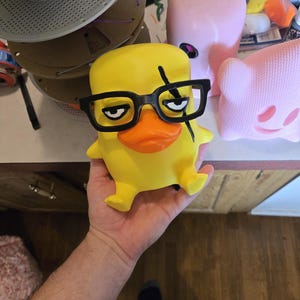May include: A yellow duck figurine with a flat top, wearing black plastic-rimmed glasses. The duck has a bright orange beak, a black scar mark over one eye, and a grumpy facial expression with narrowed eyes. The figure is smooth and matte.