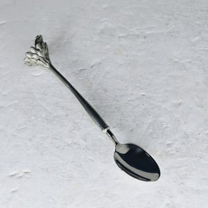 May include: A silver-colored spoon with a decorative floral design at the handle's end. The spoon has a long, slender handle and a rounded bowl. The floral design adds a touch of elegance to the utensil. The spoon is likely made of stainless steel.