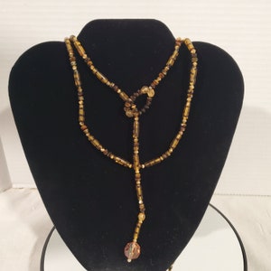 May include: A long, layered necklace made of brown and gold beads. The necklace has a teardrop-shaped pendant at the bottom.