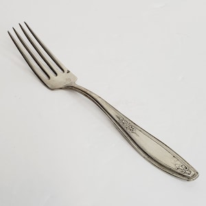 May include: A silver fork with an ornate design on the handle. The fork has four tines and a slightly curved handle.