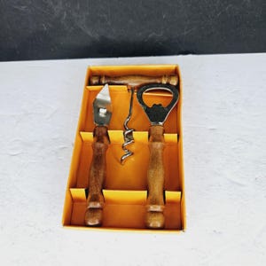 Puede incluir: Un juego de herramientas de bar vintage que incluye un abridor de botellas, un sacacorchos y un abrelatas con mangos de madera. Las herramientas se muestran en una caja de cartón amarilla con un interior marrón. Las herramientas son de metal.