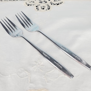 May include: Two silver forks with a floral design on the handle. The forks are on a white tablecloth.