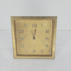 May include: A gold-toned brass clock with Roman numeral markings on the face. The clock has a simple design with a square face and a square frame.