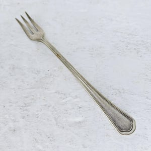 May include: A silver fork with three tines and a long, slender handle. The handle has a decorative edge. The fork is likely vintage or antique, with a slightly aged appearance.