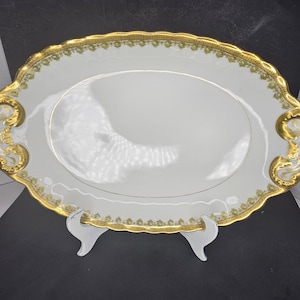 Puede incluir: Una fuente ovalada blanca con borde dorado y diseño floral verde. La fuente tiene dos asas doradas ornamentadas.