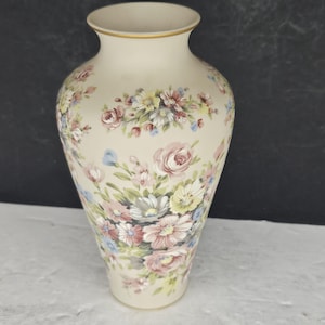 Op de afbeelding: Een crèmekleurige keramische vaas met een bloemenpatroon. De vaas heeft een gouden rand en is versierd met clusters van roze, blauwe, gele en witte bloemen. De vaas heeft een klassieke, elegante vorm.