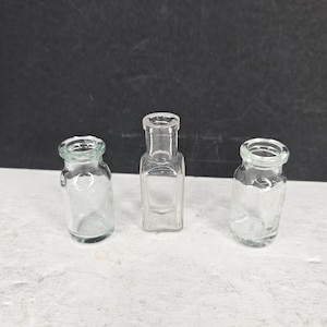 May include: Three small, clear glass bottles with open tops. The bottles are empty and have a vintage aesthetic. One bottle is rectangular, and the other two are round.