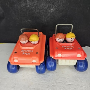 Puede incluir: Dos buggies de juguete de plástico naranja con ruedas azules. Cada buggy tiene dos pequeñas figuras en su interior, una con pelo castaño y la otra con un sombrero amarillo. El texto "Bouncing Buggy" está impreso en la parte delantera de cada juguete.