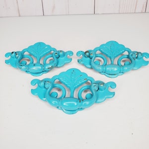 Puede incluir: Tres tiradores de cajón de metal azul turquesa con diseños ornamentados. Los tiradores tienen un acabado desgastado con acentos dorados.