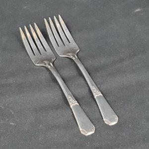 May include: Two silver forks with four tines each, arranged diagonally on a dark surface. The handles have a decorative floral pattern near the end. The forks are made of a shiny, reflective metal.
