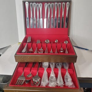 May include: A wooden box with two drawers containing a set of silverware. The top drawer has a red felt lining and the bottom drawer has a red felt lining with clear plastic sleeves. The silverware is made of stainless steel and has a polished finish.
