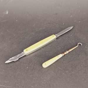May include: Two antique tools: a folding knife with a cream-colored handle and a metal blade, and a crochet hook with a cream-colored handle and a metal hook. The tools are set against a dark background.