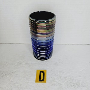 May include: A cylindrical vase with a dark interior and a metallic rim. The vase transitions from iridescent bands at the top to blue bands at the bottom. The vase has a ribbed texture.