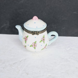 May include: A small, decorative teapot with a white base, decorated with pink floral designs and blue polka dots. The lid is light blue with blue dots and a pink knob. The teapot has a light blue handle and spout, and a bronze-coloured hinge.