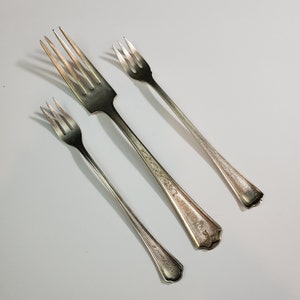 May include: Three silver forks, two are small and one is larger. The larger fork has an ornate design on the handle.