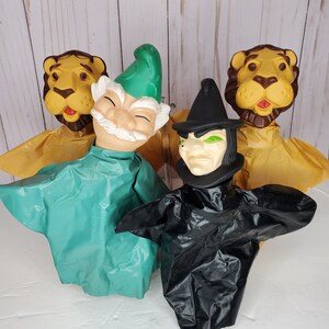 May include: Four vintage hand puppets featuring characters from The Wizard of Oz. Includes a lion, a gnome, and a wicked witch. The puppets have painted faces and fabric costumes in shades of yellow, green, and black. The puppets are displayed against a light wood background.