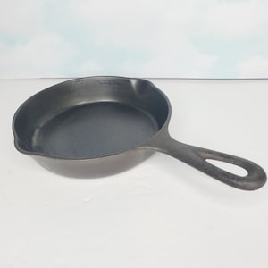 May include: A dark grey cast iron frying pan with a long handle and a rounded cooking surface. The pan is designed for stovetop or oven use. The handle has a hole for hanging. The pan is shown on a white surface.