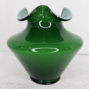 Może przedstawiać: Zielony szklany wazon z falbanką i białym brzegiem. Wazon ma błyszczące wykończenie i lekko rozszerzoną podstawę.