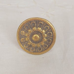 May include: A round, gold-toned metal knob with a decorative, circular design. The center of the knob is a raised, gold-toned circle.
