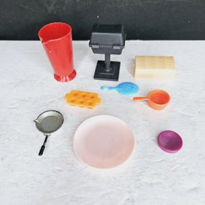 Op de afbeelding: Een verzameling miniatuurkeuken- en grillaccessoires. Inclusief een rode kan, zwarte grill, beige blok, blauwe lepel, oranje maatbeker, gele bak, zilveren pan, roze bord en paarse schijf.