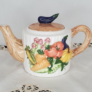 May include: A white ceramic teapot with a brown handle and a lid decorated with a variety of vegetables, including carrots, corn, tomatoes, and aubergine. The teapot is painted to look like a tree trunk.