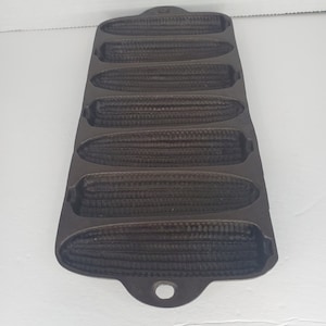 May include: A cast iron cornbread pan with six individual moulds. The pan is dark grey and has a handle on the bottom.