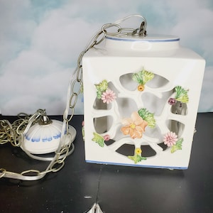 Puede incluir: Luminaria colgante de cerámica blanca con forma cuadrada y un intrincado diseño floral. La luminaria tiene una cadena y una pantalla de cerámica blanca en forma de cúpula.