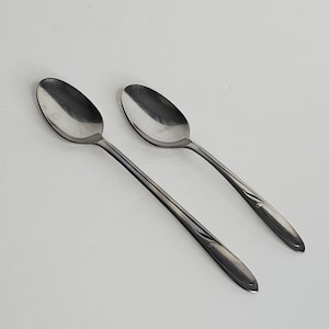 May include: Two stainless steel spoons with oval bowls and long, slender handles. The handles have a decorative, curved design near the base. The spoons are set against a plain white background, suitable for a dining or kitchen setting.