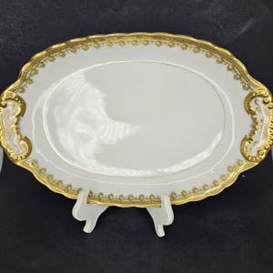 Puede incluir: Una fuente ovalada blanca con borde dorado y detalles florales verdes. La fuente tiene dos asas a los lados.