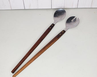 May include: A set of two wooden handled salad servers with stainless steel heads. One server has a spoon-shaped head and the other has a fork-shaped head.