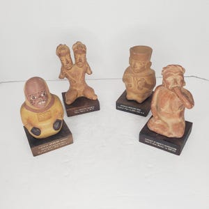 Puede incluir: Cuatro figuritas de terracota sobre bases de madera. Las figuritas representan una figura de dos cabezas, una figura con un sombrero, una figura con una capucha y una figura con barba. Cada base tiene texto que describe la figura y su origen.
