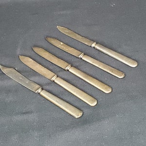 Może przedstawiać: Pięć srebrnych noży do masła ze lekko zaokrąglonymi końcówkami. Noże są ułożone w rzędzie, z uchwytami skierowanymi w prawo.