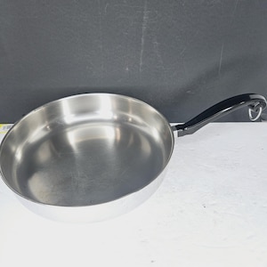 May include: A stainless steel frying pan with a black handle. The pan has a wide, shallow cooking surface and a curved rim. The handle is attached with a loop for hanging. The pan is designed for cooking on a stovetop.