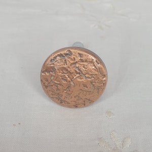 May include: A copper-colored, round cabinet knob with a textured surface. The knob has a silver-colored stem. The knob is approximately 1.5 inches in diameter.