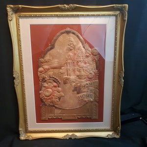 Puede incluir: Una placa de papel en relieve vintage enmarcada con una imagen en relieve de una casa y las palabras "Home Sweet Home". La placa está en un marco dorado con un passe-partout blanco.