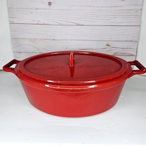 May include: A red enameled cast iron Dutch oven with a matching lid. The oval-shaped pot has two side handles and a handle on the lid. The cookware is designed for stovetop or oven use.