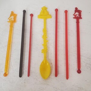 May include: Seven plastic drink stirrers with various designs.  The stirrers are yellow, orange, red, and black.  One stirrer has a spoon on the end.  The stirrers are on a white surface.