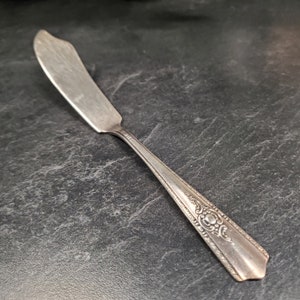 May include: A silver butter knife with an ornate, engraved handle. The knife has a slightly curved blade and a pointed tip.