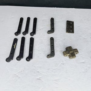 Puede incluir: Un surtido de componentes de ferretería metálicos antiguos, que incluyen bisagras y soportes. Las piezas son principalmente de color gris oscuro y bronce, con varias formas y tamaños. La ferretería parece ser para muebles o armarios.