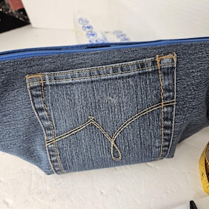 Puede incluir: Un estuche de mezclilla con cremallera azul y un bolsillo trasero. El bolsillo tiene costuras decorativas en un color beige contrastante. El estuche está hecho de material de mezclilla azul.