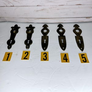 May include: Five antique brass keyhole escutcheon plates arranged in a row, each featuring a distinct elongated shape with a circular top opening and a lower keyhole. Each plate is labeled with a small yellow square numbered one through five.