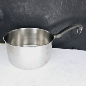 May include: A stainless steel saucepan with a black handle. The pot has a rounded body and a single, curved handle. The handle is attached to the side of the pot and has a loop at the end. The pot is sitting on a white surface.