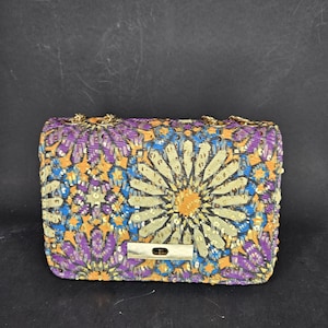 May include: A small, purple, blue, and gold patterned purse with a gold chain strap and a gold clasp.