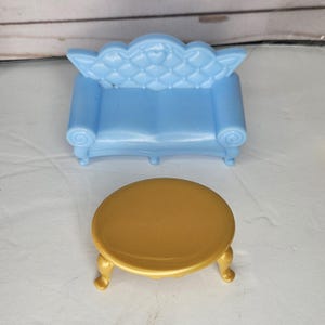 Op de afbeelding: Een miniatuur lichtblauwe bank met een gecapitonneerde rugleuning en een goudkleurige ronde tafel met drie gebogen poten. De meubelset is ontworpen voor een poppenhuis of vergelijkbaar speelgoed.