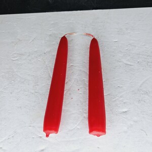 May include: Two bright red taper candles, standing upright against a white textured surface. The candles are slightly melted at the bottom. The wicks are visible at the top. The background is black.
