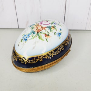May include: A porcelain egg-shaped trinket box with a blue and gold trim. The lid is decorated with hand-painted floral designs in shades of pink, blue, yellow, and green.
